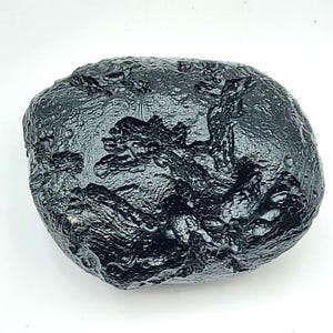May include: A dark, irregularly shaped, black stone with a textured surface. The stone has a glossy sheen and appears to be a natural formation. The surface is covered in ridges and small, rounded bumps. The overall shape is oval.