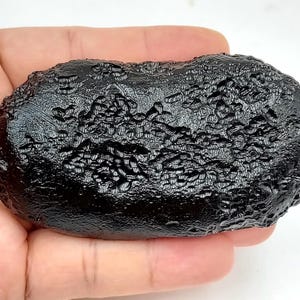 May include: A dark, irregularly shaped rock with a textured surface. The rock is held in a hand, and appears to be a meteorite. The colour is a deep black, and the surface has a rough, pitted appearance.
