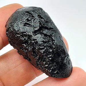 May include: A dark, glossy black tektite with a rough, textured surface. The elongated, teardrop-like shape of the natural stone is visible. The tektite is held in a person's hand.