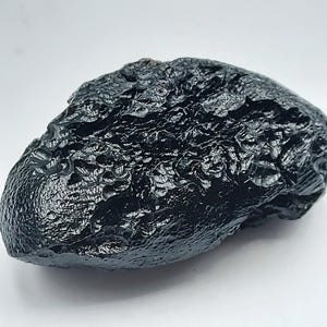 May include: A dark, glossy, black tektite specimen with a textured, uneven surface. The natural glass rock has a rounded, irregular shape and appears to be a meteorite. The background is plain white.
