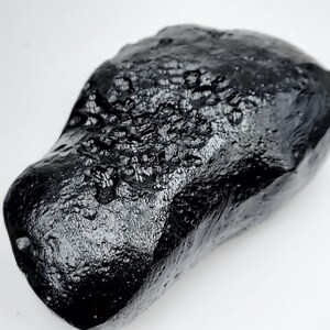 May include: A dark, irregularly shaped, black stone with a glossy, textured surface. The stone has a rough, pitted appearance, with areas of raised and uneven texture. The overall shape is rounded and organic, with a smooth, reflective sheen.