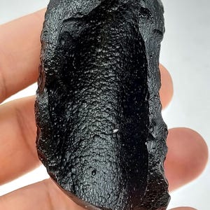 May include: A dark, irregularly shaped, black stone with a textured surface. The stone has a deep, concave area on one side, creating a unique, organic form. The overall appearance is rough and natural, with a glossy sheen.