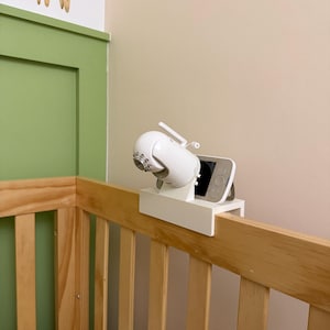May include: A white baby monitor with a screen and antenna is mounted on a white bracket attached to a wooden crib. The crib is made of light-colored wood. A giraffe illustration is on the wall in the background.