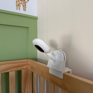 May include: A white baby monitor camera is attached to the wooden railing of a crib. The camera has a curved design and is positioned to face the interior of the crib. The crib is made of light-colored wood.