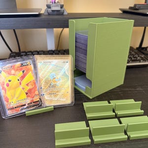 May include: A green card holder with a stack of cards inside, alongside individual card dividers. Two trading cards are visible, one featuring Pikachu and the other Squirtle. The holder and dividers are made of a solid, matte green material.