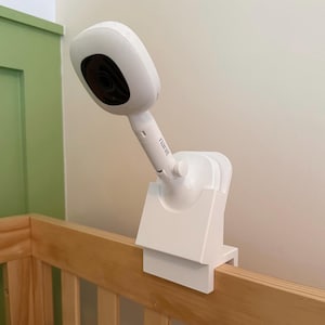 May include: A white Nanit baby monitor camera mounted on a white plastic clamp attached to the top rail of a wooden crib. The camera features a rounded head with a black lens and a cylindrical neck with the brand name Nanit printed on it.