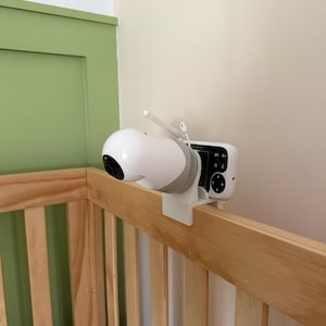 May include: A white and gray baby monitor camera and receiver attached to a wooden crib. The camera has a rounded design with a black lens and two antennas. The receiver has a small screen and control buttons. The crib is made of light-colored wood.