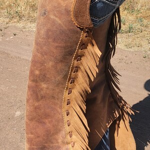 May include: Brown leather chaps with decorative stitching and fringe. The chaps have a suede-like texture on the upper portion and are worn over blue jeans. The chaps are designed for horseback riding and western wear.