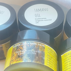 May include: Close-up of several glass jars with black lids. One jar is labeled "TAMANU OIL" with "frankincense, tangerine essential oils" below. Another jar is labeled "SEASIDE GOLDENROD SALVE" with "Herbal Scent with Essential Oils".