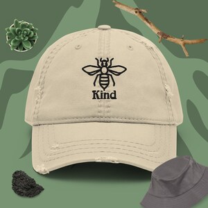 Op de afbeelding: Een beige, verweerde baseballpet met een zwarte geborduurde bij en het woord "Kind" eronder.