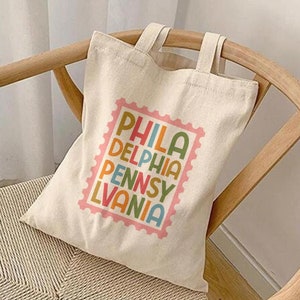 May include: A white canvas tote bag with a colorful graphic of a stamp that says "PHILA DELPHIA PENNSY LVANIA". The bag is sitting on a wooden chair.