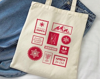 May include: A white canvas tote bag with red printed designs. The designs include a snowflake, a mountain range, ski poles, a cocktail glass, and the text "With love from Aspen", "Amanda's Bachelorette Weekend", "Experts Only", and "Apres Ski Club".