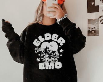 May include: A black sweatshirt with a white graphic print of a cartoon character with the text "Elder Emo" and the words "It's not a phase" above the character. The model is wearing the sweatshirt with cowboy boots.