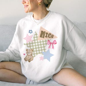 May include: White sweatshirt with a patchwork design featuring a teddy bear, stars, a pink bow, and the word "Nicu" on a banner. The patchwork includes gingham and floral patterns in pastel colors.