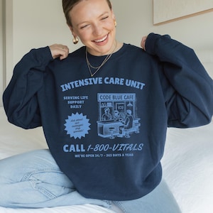 May include: Navy blue sweatshirt with light blue graphic text and illustrations. The text reads "INTENSIVE CARE UNIT" and "CODE BLUE CAFE". Additional text includes "SERVING LIFE SUPPORT DAILY" and "CALL 1-800-VITALS". The sweatshirt is a long-sleeved crew neck.