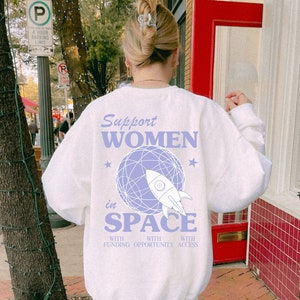 May include: A white sweatshirt with a purple graphic of a rocket ship in space. The text on the sweatshirt reads "Support Women in Space with Funding Opportunity Access".