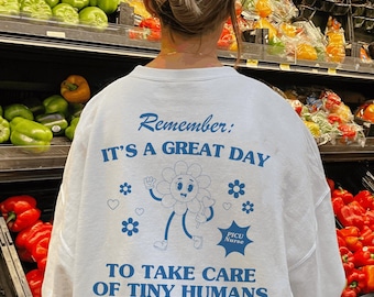 May include: A white long-sleeved shirt with a blue graphic of a smiling flower with the text "Remember: It's a great day to take care of tiny humans."