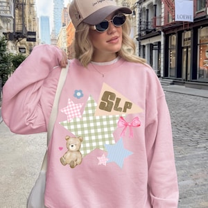 May include: A light pink sweatshirt with a whimsical design featuring stars, a teddy bear, and the letters "SLP." The design incorporates various patterns and a pink bow. The person is wearing a baseball cap.