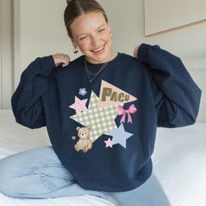 May include: Navy blue sweatshirt with a colorful graphic design. The design includes a teddy bear, stars, a pink bow, and the word "PACU" on a geometric shape. The sweatshirt is a long-sleeved crew neck.