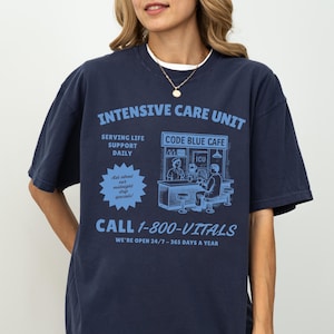 May include: Navy blue t-shirt with the text "INTENSIVE CARE UNIT" and "CALL 1-800-VITALS" in light blue. The shirt features a graphic of a cafe scene and the words "SERVING LIFE SUPPORT DAILY".
