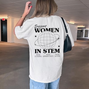 May include: White t-shirt with the text "Support Women in STEM" in black, featuring a globe design. The shirt also includes the phrases "with funding, opportunity, access." The person is wearing light blue jeans and white sneakers.