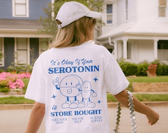 May include: White t-shirt with a blue graphic of a cartoon pill with the text "It's Okay If Your Serotonin is Store Bought" and the phrases "Take Your Meds", "Ask For Help", and "Be Kind Always".