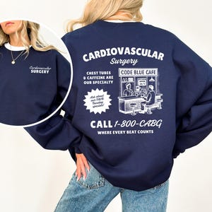 May include: Navy blue sweatshirt with white text that reads "Cardiovascular Surgery" on the front and back. The back also features a graphic of a cafe and the text "Chest tubes & caffeine are our specialty." The sweatshirt has a white collar.