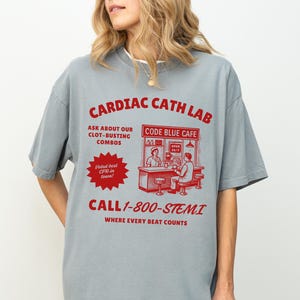 May include: A gray t-shirt with a red graphic that reads "CARDIAC CATH LAB" and "CALL 1-800-STEMI". The graphic includes text about "CLOT-BUSTING COMBOS" and "CODE BLUE CAFE". The shirt has short sleeves.