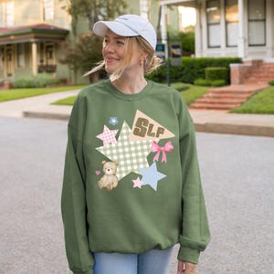K&ouml;nnte beinhalten: Olivgr&uuml;nes Sweatshirt mit farbenfrohem Grafikdesign. Das Design umfasst Sterne, einen Teddyb&auml;ren, eine Schleife und die Buchstaben "SLP". Das Sweatshirt ist ein l&auml;ssiger, bequemer Stil.