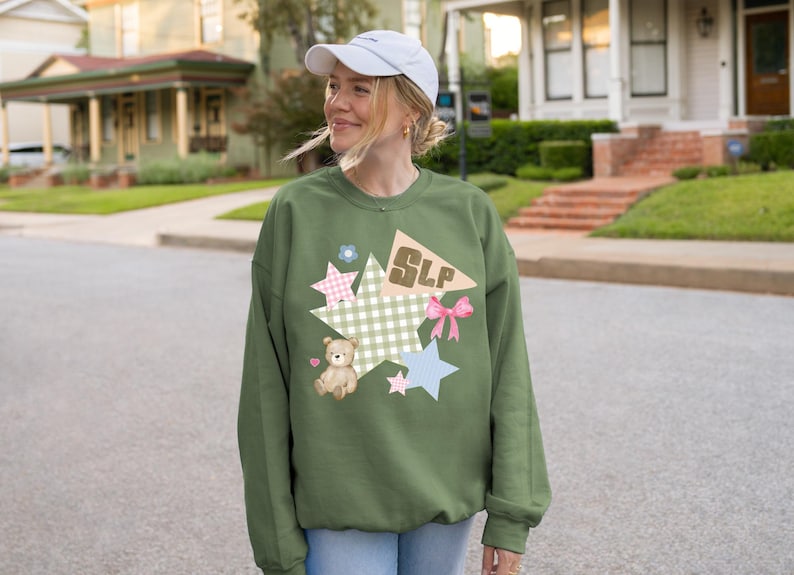 K&ouml;nnte beinhalten: Olivgr&uuml;nes Sweatshirt mit farbenfrohem Grafikdesign. Das Design umfasst Sterne, einen Teddyb&auml;ren, eine Schleife und die Buchstaben "SLP". Das Sweatshirt ist ein l&auml;ssiger, bequemer Stil.