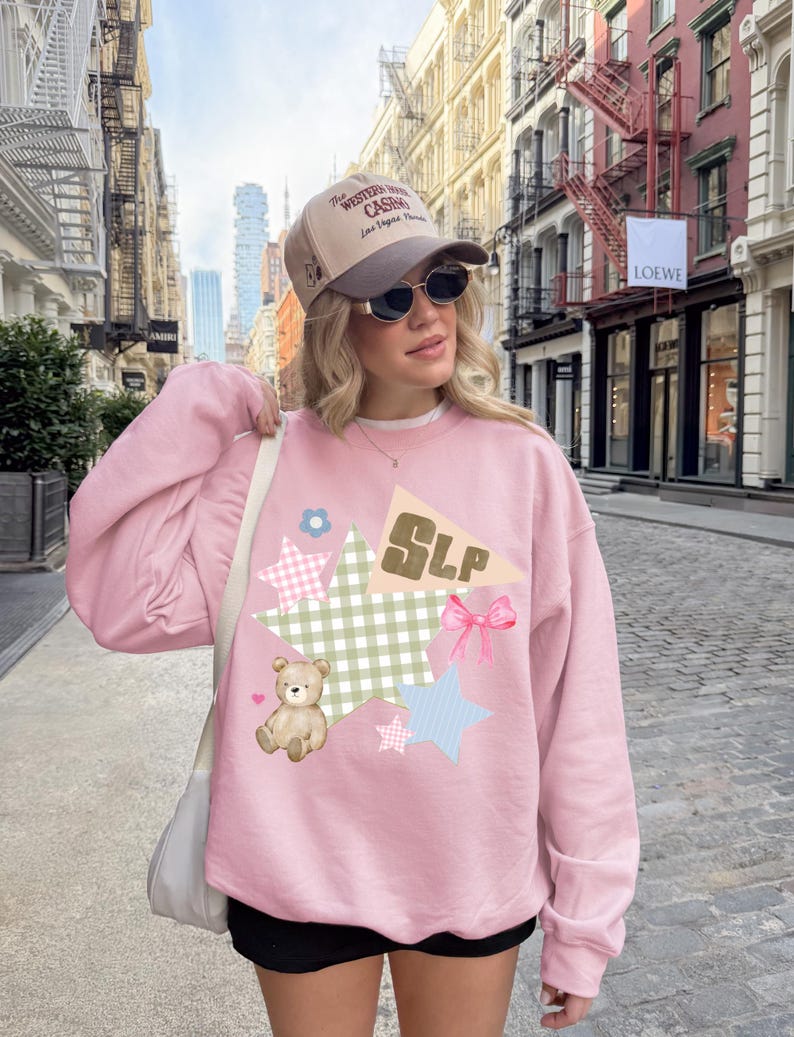 K&ouml;nnte beinhalten: Ein hellrosa Sweatshirt mit einem verspielten Design mit Sternen, einem Teddyb&auml;r und den Buchstaben "SLP". Das Design beinhaltet verschiedene Muster und eine rosa Schleife. Die Person tr&auml;gt eine Baseballkappe.
