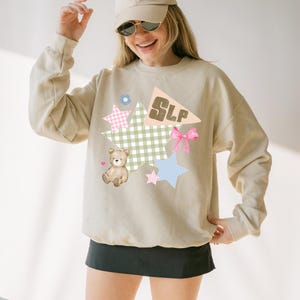 K&ouml;nnte beinhalten: Beiges Sweatshirt mit einem Grafikdesign mit einem Teddyb&auml;ren, Sternen, einer Schleife und den Buchstaben "SLP" auf einer Wimpel. Das Design umfasst Pastellfarben und ein Vichy-Muster. Die Person tr&auml;gt eine beige Kappe und Sonnenbrille.