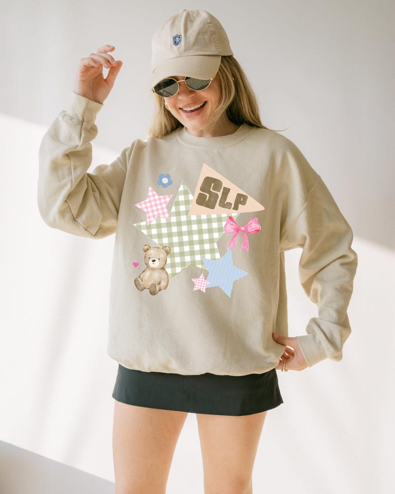K&ouml;nnte beinhalten: Beiges Sweatshirt mit einem Grafikdesign mit einem Teddyb&auml;ren, Sternen, einer Schleife und den Buchstaben "SLP" auf einer Wimpel. Das Design umfasst Pastellfarben und ein Vichy-Muster. Die Person tr&auml;gt eine beige Kappe und Sonnenbrille.
