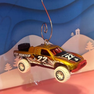 May include: A gold and white toy truck with a red bumper and the words "HW Winter 22" on the side. The truck is hanging from a silver hook and is a Christmas ornament.