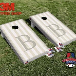 Könnte beinhalten: Zwei Cornhole-Boards mit einem gestreiften Design und dem Buchstaben "B" in Grau. Die Boards haben ein Loch oben und werden von Beanbags begleitet. Die Boards befinden sich auf einer grünen, grasbewachsenen Oberfläche. Das Logo "Cornhole Depot" ist sichtbar.