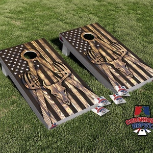 Könnte beinhalten: Zwei Holz-Cornhole-Bretter mit einem verwitterten Holzlook und einem amerikanischen Flaggen-Design. Die Bretter zeigen eine Hirschschädelgrafik mit Geweih. Die Bretter sind für ein Cornhole-Spiel aufgebaut, mit Säcken auf dem Boden.