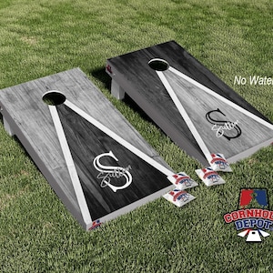 Könnte beinhalten: Zwei Holz-Cornhole-Bretter mit einem schwarzen und weißen Holzmaserungsmuster. Jedes Brett hat ein weißes Dreieck mit schwarzem Umriss und einen weißen Buchstaben "S" mit dem Namen "Sutton" in Kursivschrift. Die Bretter befinden sich auf einer grünen Grasfläche mit vier Bohnensäcken im Vordergrund.