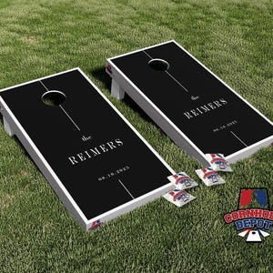 Könnte beinhalten: Zwei schwarze Cornhole-Boards mit weißen Rändern und Beinen werden auf grünem Gras gezeigt. Jedes Board hat ein zentriertes Loch und den Text "the REIMERS" und "08.16.2025". Vier Beanbags und ein Logo sind ebenfalls sichtbar.