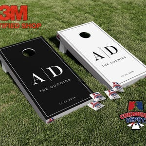 Könnte beinhalten: Zwei personalisierte Cornhole-Boards, eines in Schwarz und eines in Weiß, mit den Initialen "AD" und dem Namen "THE GODWINS" aufgedruckt. Das Datum "10.09.2026" ist ebenfalls auf den Boards aufgedruckt. Vier Beanbags sind sichtbar.