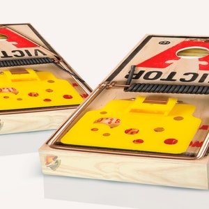Op de afbeelding: Twee houten spelborden in de vorm van muizenval met een geel kaasvormig doel en het woord "VICTOR" gedrukt op de borden.