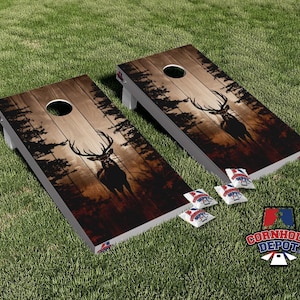 Puede incluir: Dos tableros de cornhole de madera con un diseño de veta de madera rústica y una silueta de un ciervo en un entorno de bosque. Los tableros están configurados para un juego de cornhole, con bolsas de frijoles en primer plano.