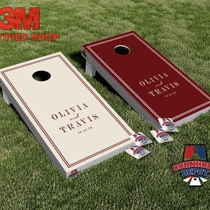 Op de afbeelding: Twee gepersonaliseerde cornhole-boards, één crèmekleurig en één bordeauxrood, met de namen "Olivia en Travis" en de datum "10.24.26" erop gedrukt. De boards staan op een groene grasmat, met zitzakken en een logo.