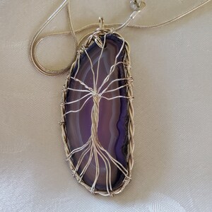 May include: A silver necklace featuring a pendant made from a polished, purple and white agate slice. The agate is encased in silver wire, shaped into a tree design. The necklace has a silver chain and clasp.