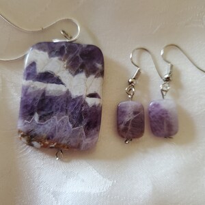 May include: A set of amethyst jewelry featuring a pendant and earrings. The pendant is a large, rectangular piece of amethyst with a swirling pattern of purple and white. The earrings are smaller, rectangular pieces of amethyst with a similar swirling pattern.