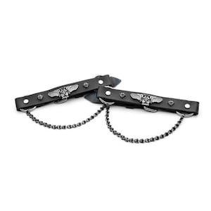 Puede incluir: Un par de correas de botas de moto de cuero negro con detalles metálicos plateados. Cada correa tiene un diseño de cruz alada y una cadena unida a los anillos metálicos.