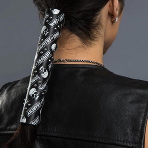 May include: Black leather ponytail holder with a skull and crossbones pattern and the text "Sons of Anarchy" repeated. The holder is studded with silver-toned metal studs.