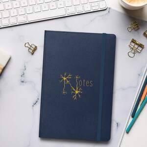 May include: A dark blue notebook with a gold neuron graphic and the word "Notes" on the cover. The notebook is on a white marble surface with a keyboard, pencils, and paper clips.