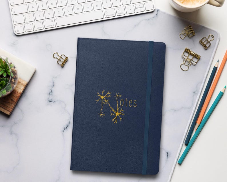 May include: A dark blue notebook with a gold neuron graphic and the word "Notes" on the cover. The notebook is on a white marble surface with a keyboard, pencils, and paper clips.