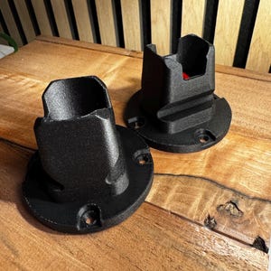 Op de afbeelding: Twee zwarte kunststof wandbeugels met een vierkante opening aan de bovenkant. De beugels zijn ontworpen om een apparaat of object te bevestigen.