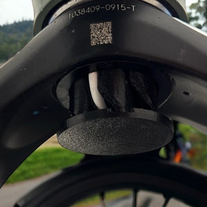 Peut inclure: Gros plan sur une fourche de vélo noire avec un câble blanc qui la traverse. La fourche est recouverte d'un cache en plastique noir avec une surface texturée et un code QR avec le numéro 1038409-0915-T.